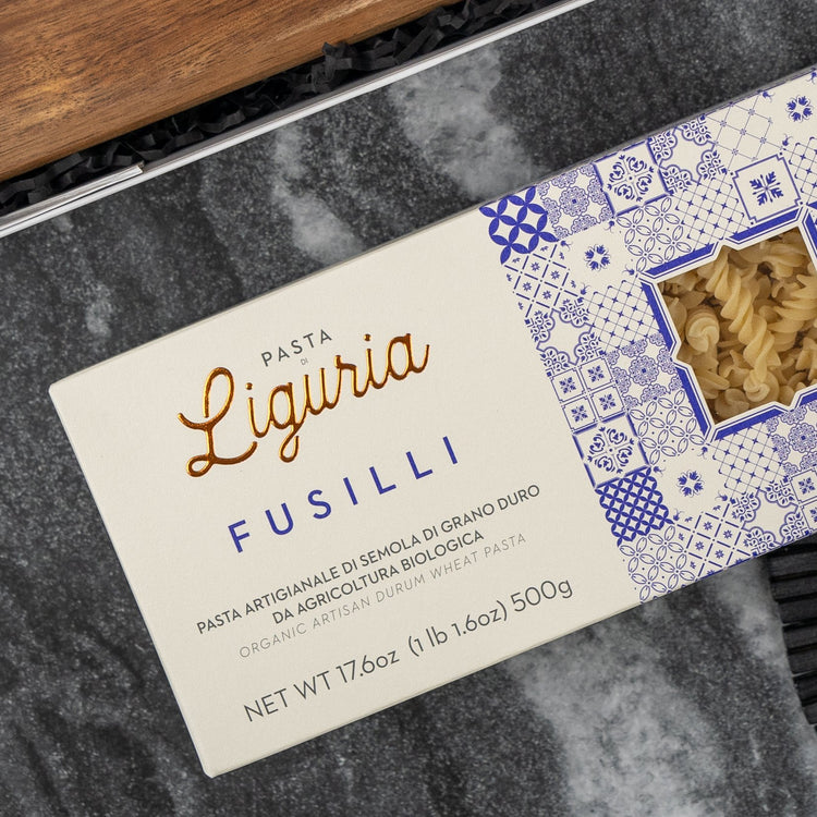 A box from The Hamper Boutique Co’s Italian Kitchen Hamper is displayed on a dark, textured surface. The packaging is white with blue patterns and a clear window, with the label in both Italian and English.