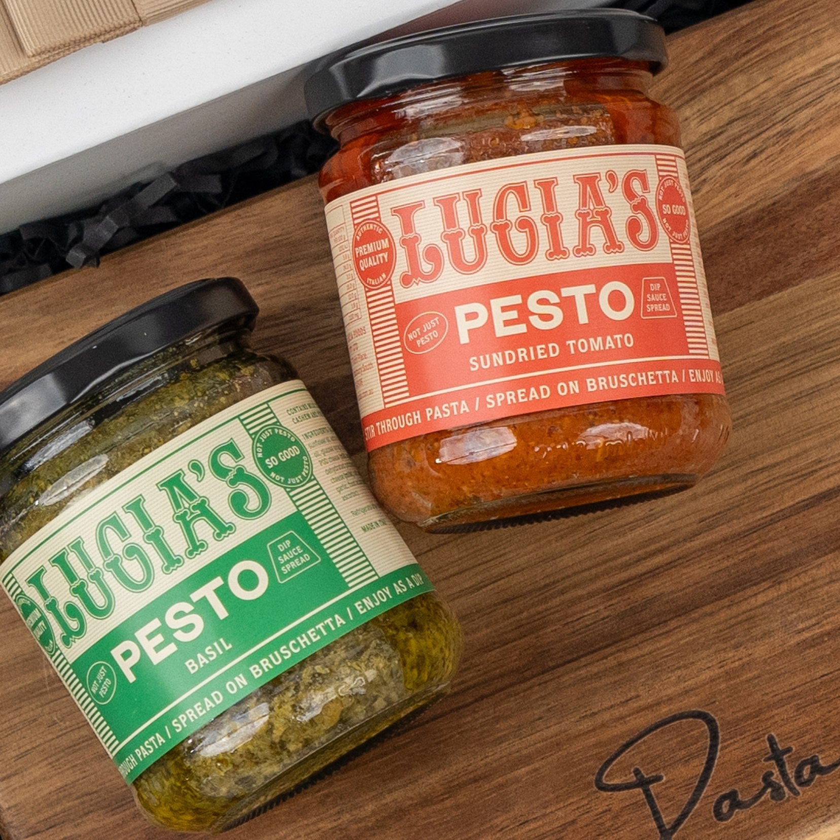 Two jars from The Hamper Boutique Co's Italian Kitchen Hamper—one basil pesto, one sundried tomato pesto—sit on a wooden surface, with “Pasta” written on the board.