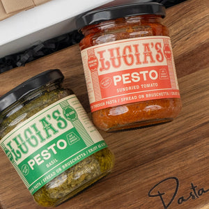 Two jars from The Hamper Boutique Co's Italian Kitchen Hamper—one basil pesto, one sundried tomato pesto—sit on a wooden surface, with “Pasta” written on the board.