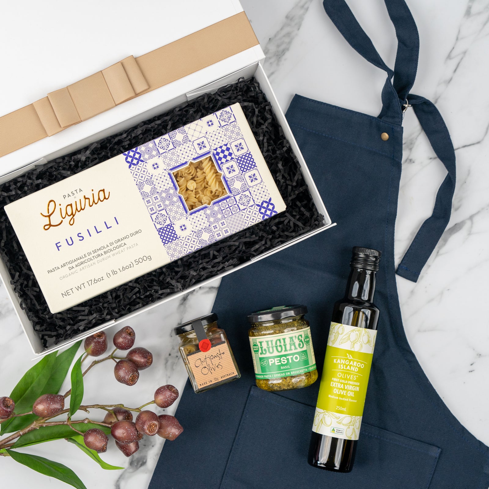 Flat lay of The Hamper Boutique Co's Cucina Essentials Hamper with blue apron, Liguria fusilli, calamata olives, pesto jar, extra virgin olive oil bottle, and olive leaves on a marble surface.