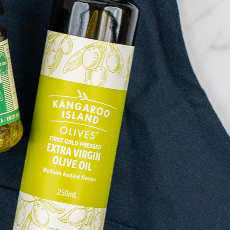A bottle of The Hamper Boutique Co's Cucina Essentials Hamper extra virgin olive oil with a green and white label rests on a dark blue cloth, beside the edge of another glass jar product.
