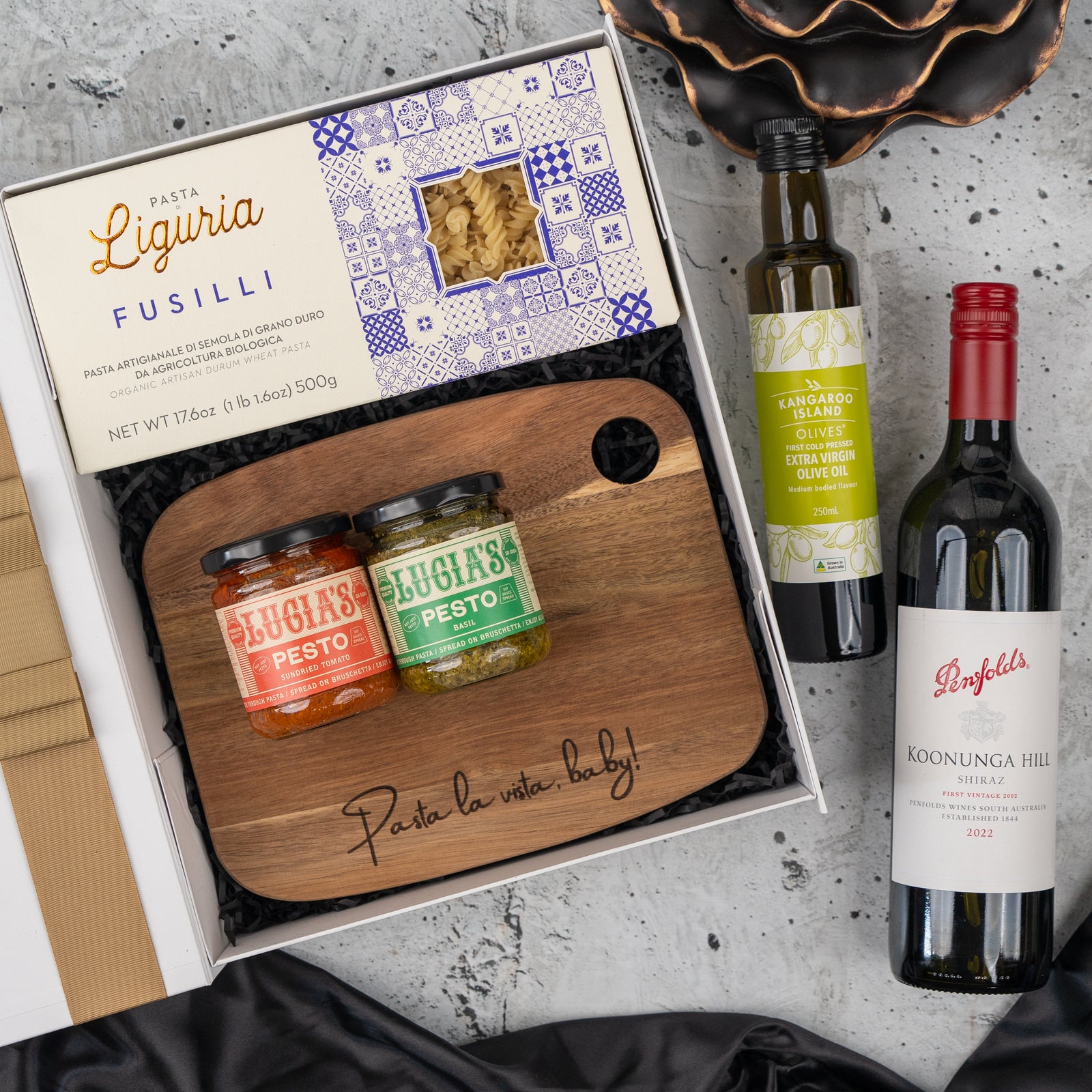The Hamper Boutique Co’s Italian Feast and Wine Hamper includes pasta, two pesto jars, olives, red wine, and a wooden board engraved with "Pasta la vista, baby!", all beautifully arranged on a stone surface.