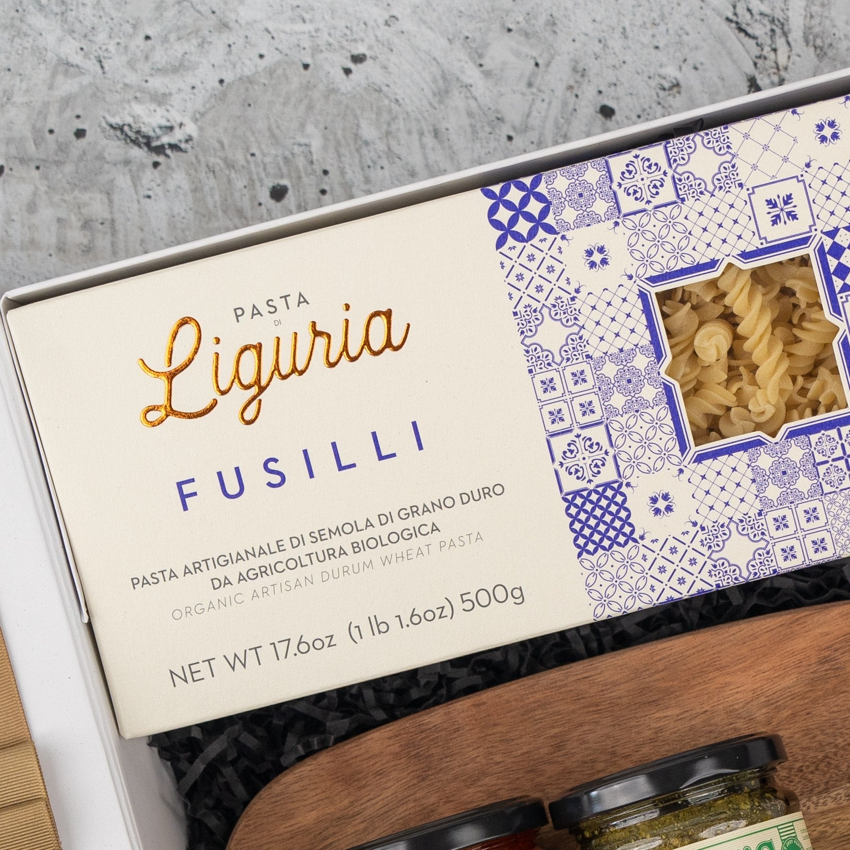 A close-up of The Hamper Boutique Co’s Italian Feast and Wine Hamper shows a box of Liguria Fusilli pasta with a blue and white design, partially opened to reveal the pasta, nestled among jars and black shredded paper in the gift set.