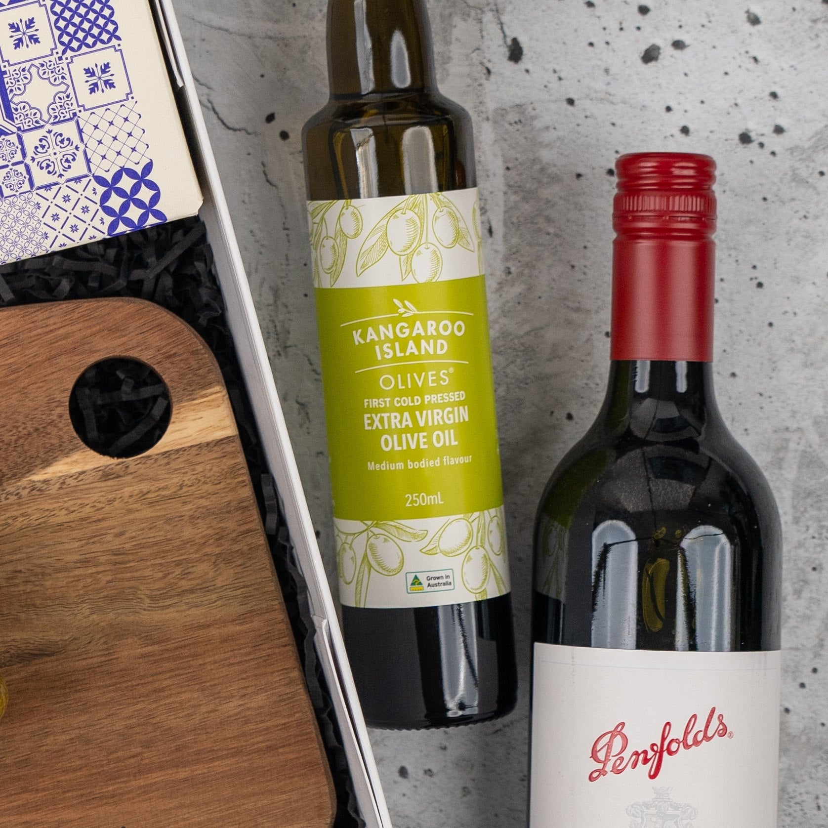 The Hamper Boutique Co's Italian Feast and Wine Hamper, featuring premium wine and gourmet treats, is arranged in a stylish gift basket with a wooden board and blue-and-white packaged item on a gray surface.