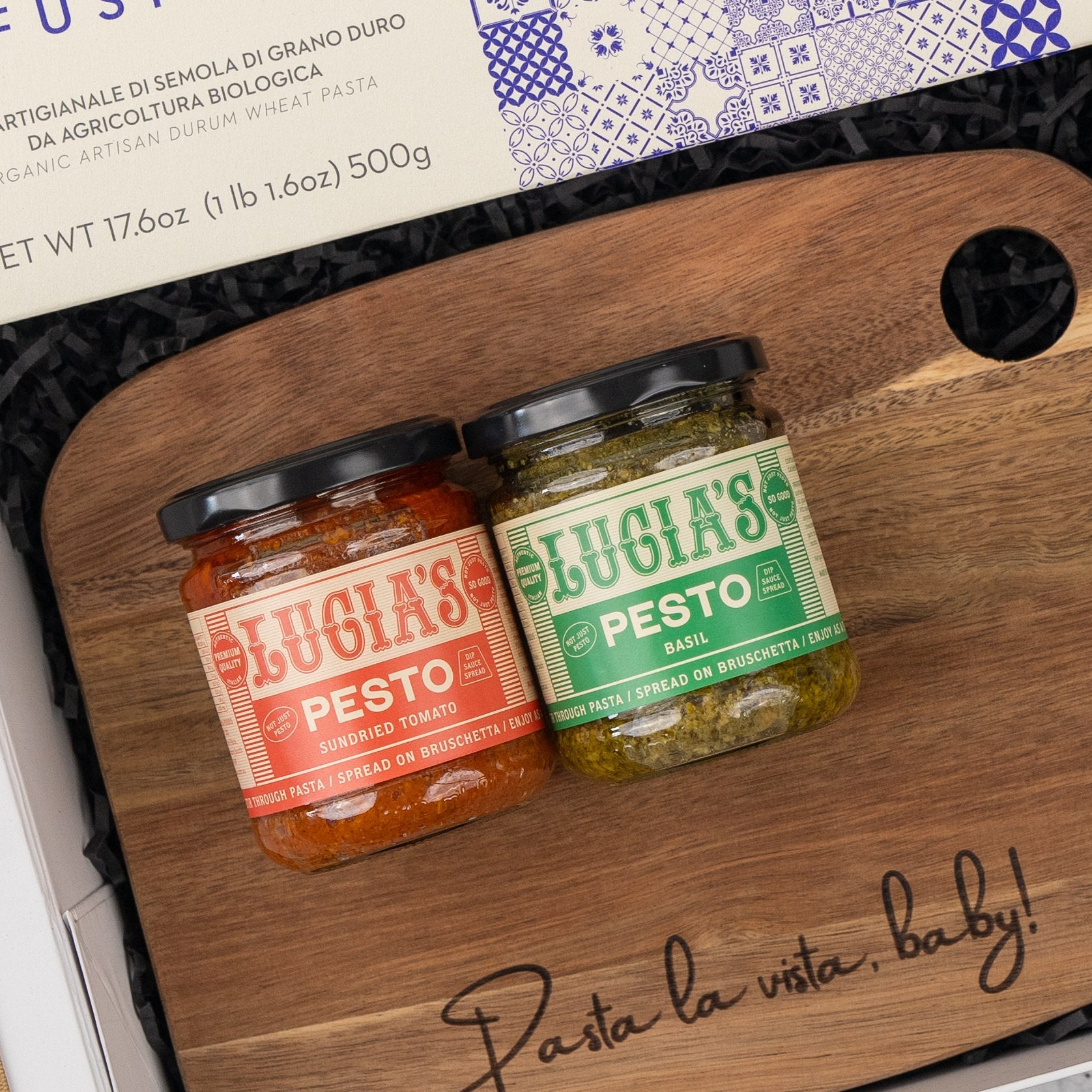 The Italian Feast and Wine Hamper by The Hamper Boutique Co features jars of sundried tomato and basil pesto side by side on a wooden board engraved with “Pasta la vista, baby!”, placed next to a box of pasta.
