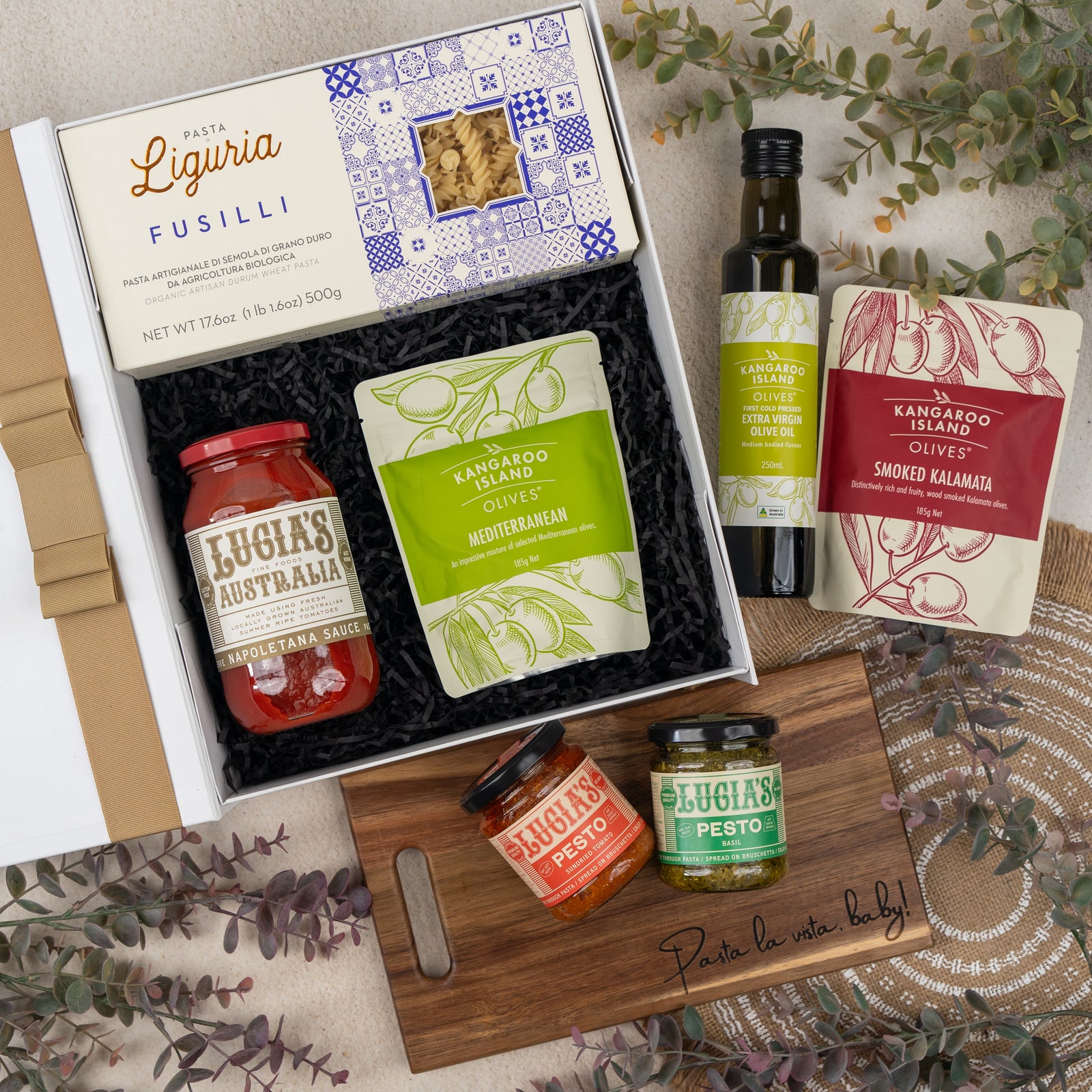 The Hamper Boutique Co's Mediterranean Pantry Hamper features pasta, sauce, olive oil, olives, two pesto jars, and a “Pasta la vista baby” wooden board, beautifully arranged with a decorative leaf on a textured surface.