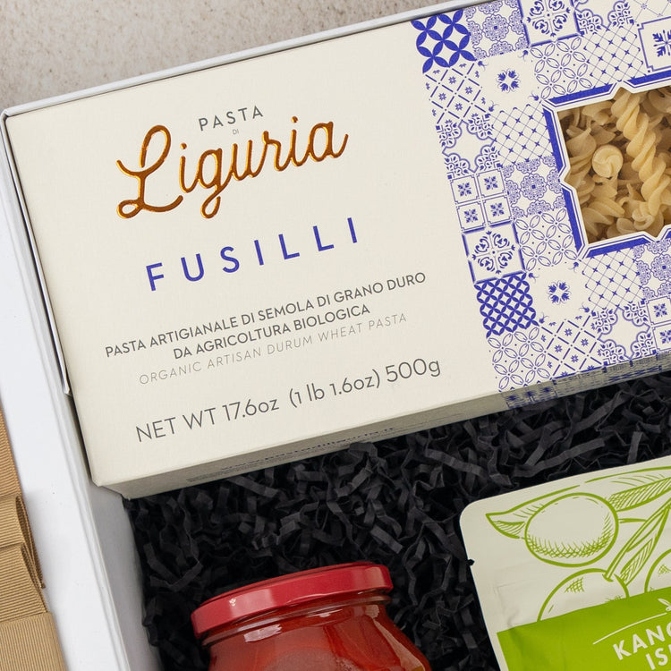 The Mediterranean Pantry Hamper by The Hamper Boutique Co features a box of Liguria fusilli pasta with a blue and white design, nestled in black shredded paper, alongside a jar of red sauce and a green-labeled package.