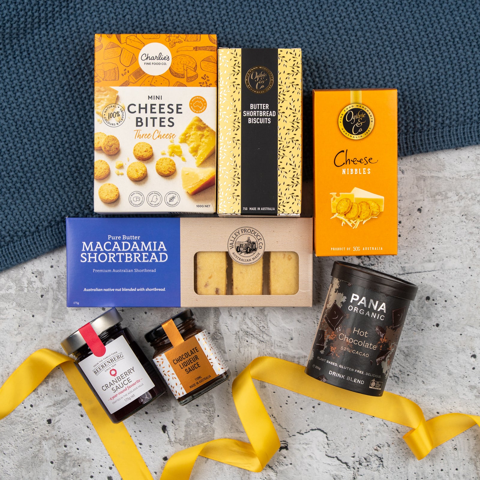 The Hamper Boutique Co’s Sweet and Savoury Delights Hamper features cheese bites, biscuits, shortbread, chocolate sauce, cranberry chutney, and hot chocolate on marble with a yellow ribbon and blue fabric.