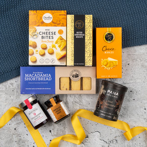 The Hamper Boutique Co’s Sweet and Savoury Delights Hamper features cheese bites, biscuits, shortbread, chocolate sauce, cranberry chutney, and hot chocolate on marble with a yellow ribbon and blue fabric.