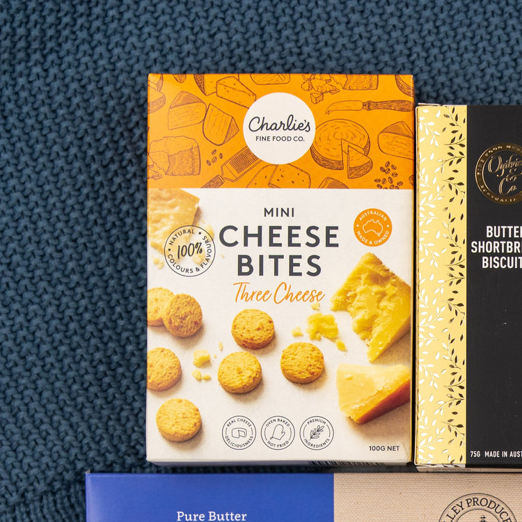 A box of The Hamper Boutique Co Sweet and Savoury Delights Hamper sits on a blue textured surface, with packaging featuring images of assorted treats.