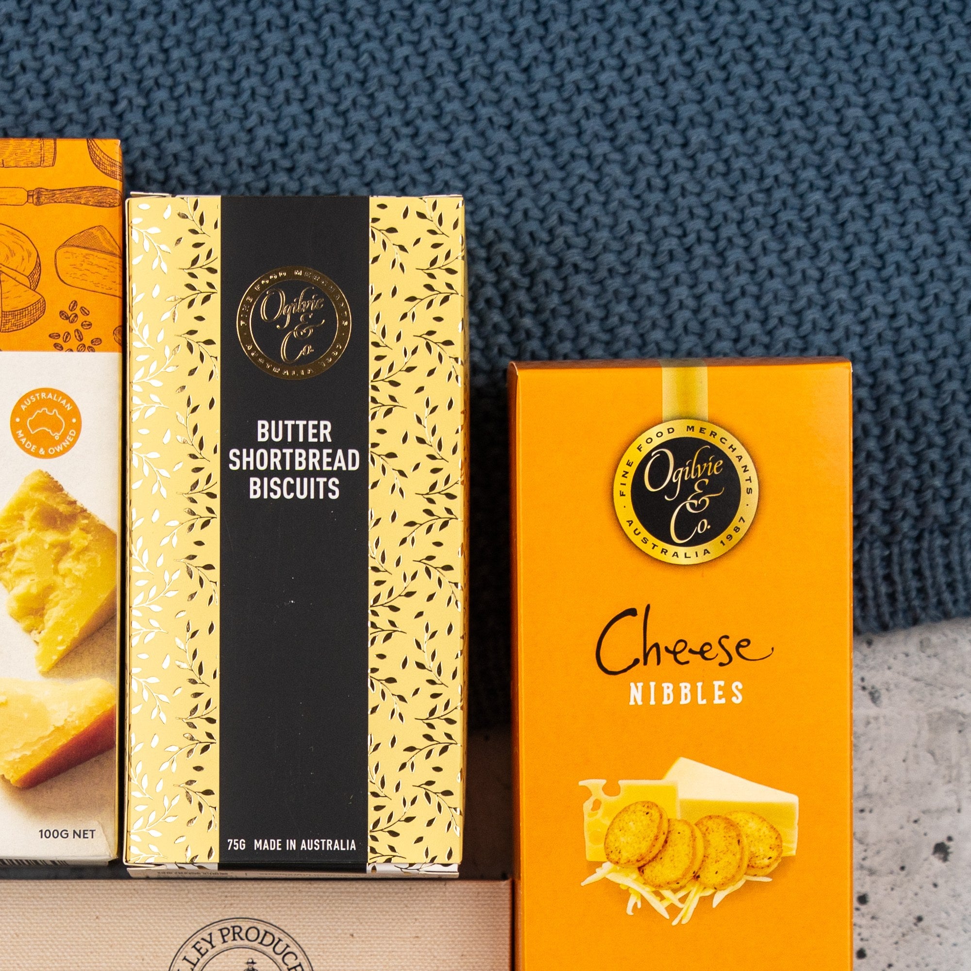 The Hamper Boutique Co Sweet and Savoury Delights Hamper features a selection of boxed snacks displayed on a blue textured surface, including butter shortbread biscuits, cheese nibbles, and crackers.