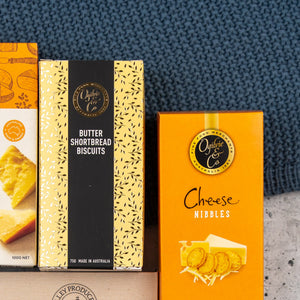The Hamper Boutique Co Sweet and Savoury Delights Hamper features a selection of boxed snacks displayed on a blue textured surface, including butter shortbread biscuits, cheese nibbles, and crackers.