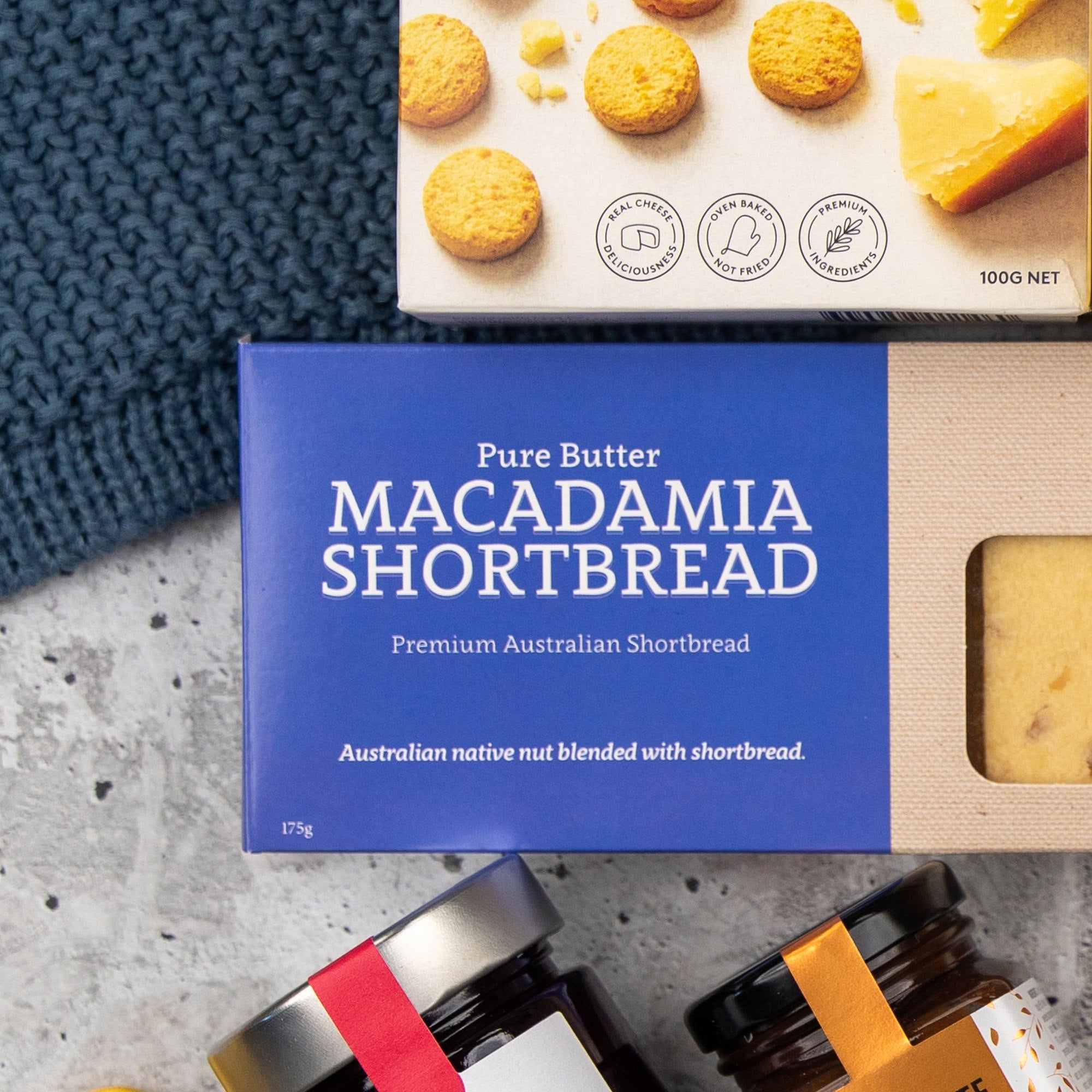 A Sweet and Savoury Delights Hamper by The Hamper Boutique Co sits on a surface, with shortbread cookies pictured above and jars with red ribbons partially visible in the corner.