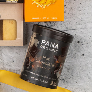 A Sweet and Savoury Delights Hamper from The Hamper Boutique Co is displayed on a gray surface, featuring a Pana Organic Hot Chocolate blend, a yellow ribbon, biscuits, and an orange box partially visible at the top.