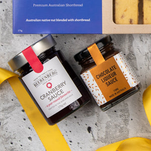 Two jars of Cranberry Sauce and Chocolate Liqueur Sauce from The Hamper Boutique Co's Sweet and Savoury Delights Hamper rest on a gray surface beside a blue shortbread box, yellow ribbon, and a partially visible shortbread biscuit.