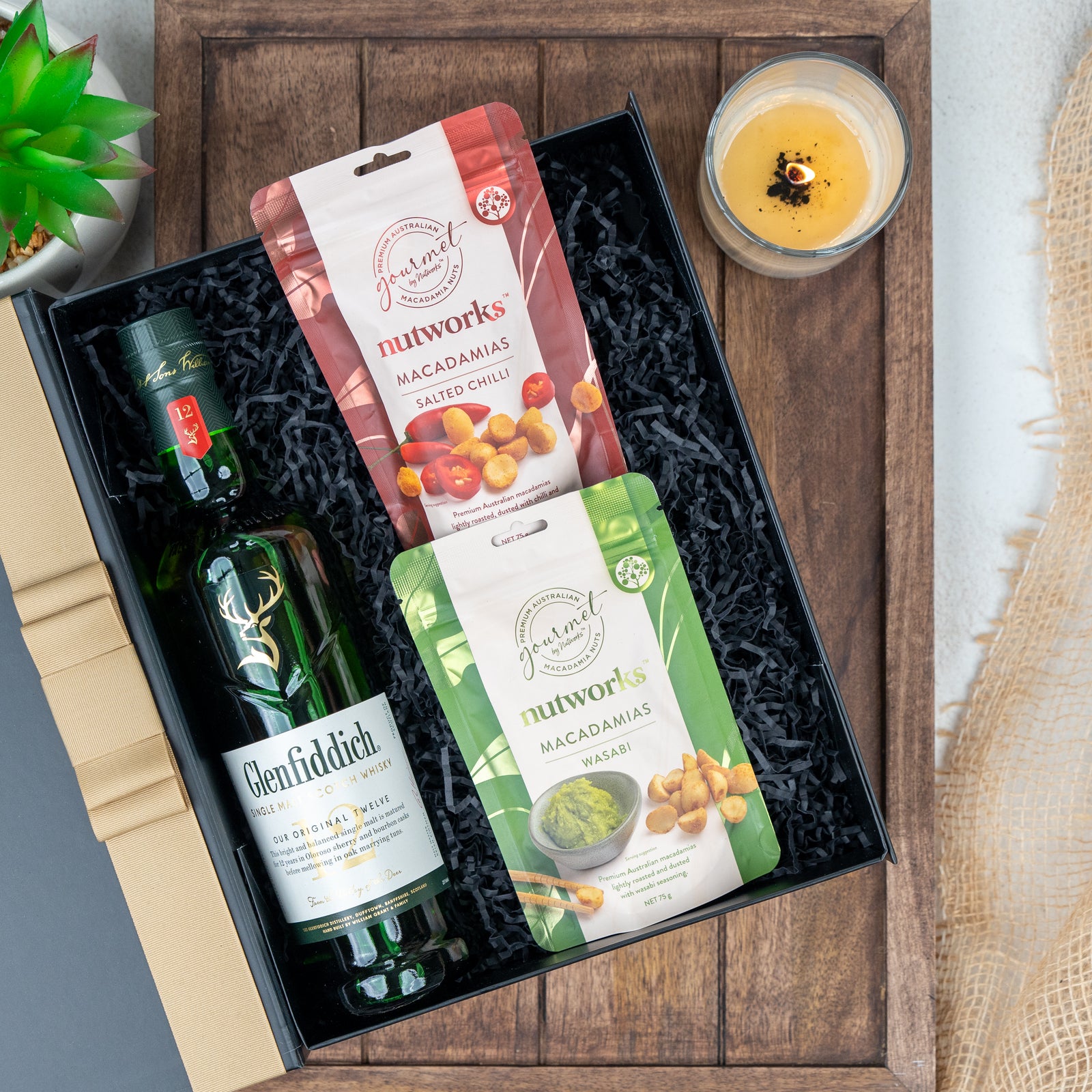 The Hamper Boutique Co's Glenfiddich Tasting Hamper includes Glenfiddich Scotch whisky, Nutworks salted chili and wasabi macadamia nut packs, displayed on black shredded paper, plus a candle and a small potted plant.