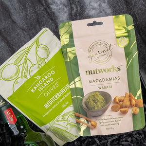 The Glenfiddich Explorer’s Hamper by The Hamper Boutique Co features two snack packs: Mediterranean olives in a green pouch with olive illustrations and Wasabi Macadamias in a beige pouch with wasabi and macadamia nut images.