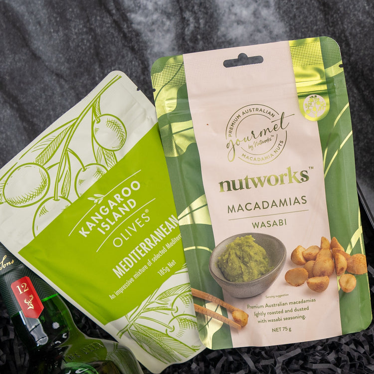 The Glenfiddich Explorer’s Hamper by The Hamper Boutique Co features two snack packs: Mediterranean olives in a green pouch with olive illustrations and Wasabi Macadamias in a beige pouch with wasabi and macadamia nut images.