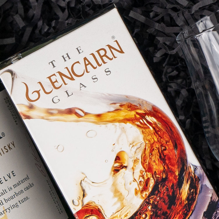 A box labeled Glenfiddich Explorer's Hamper from The Hamper Boutique Co sits beside a clear whisky glass on a dark textured surface.