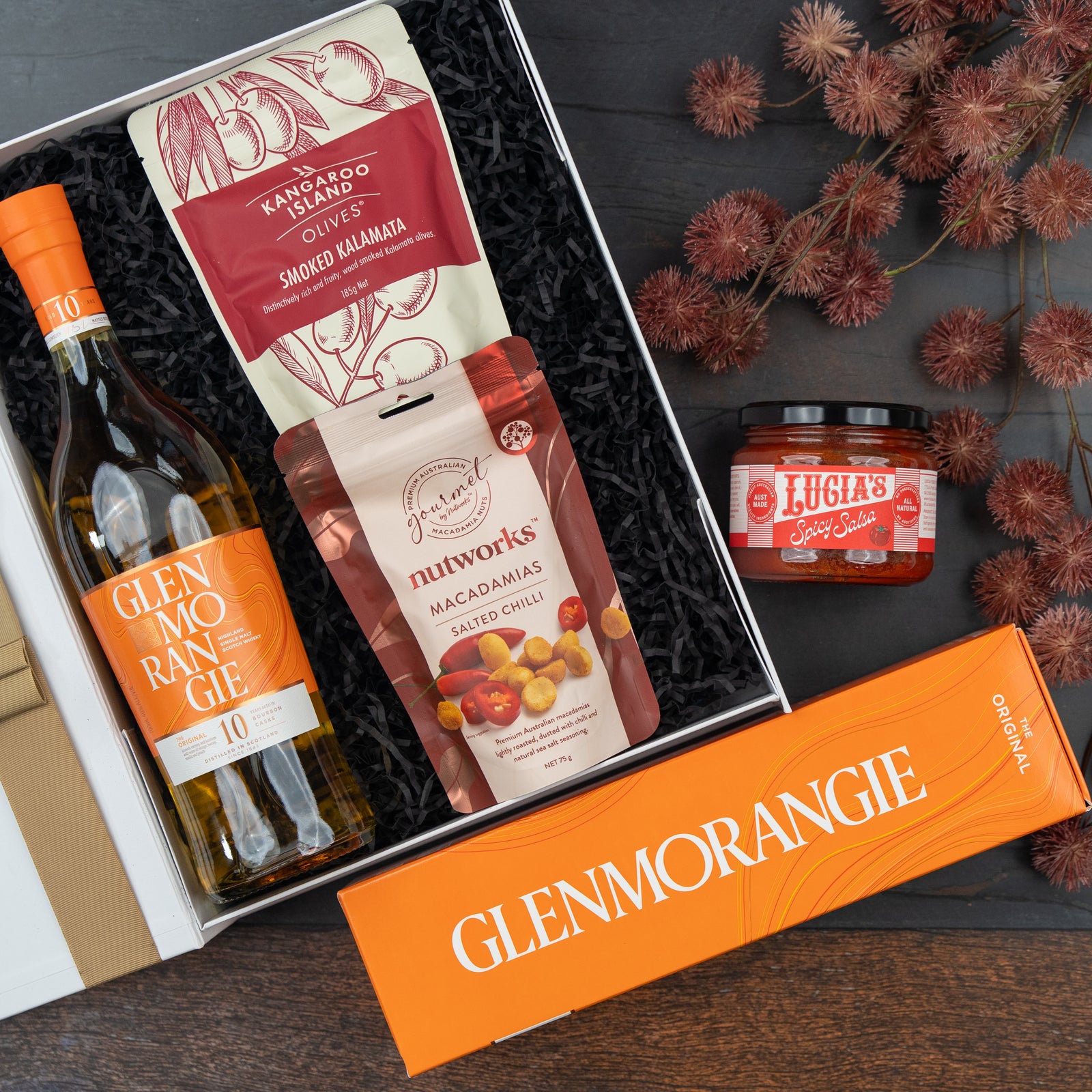 The Hamper Boutique Co’s Glenmorangie Moments Hamper features a bottle of Glenmorangie whisky, smoked Kalamata olives, salted chili macadamias, and Lucias spicy salsa, elegantly arranged on a dark wood surface with dried brown flowers.