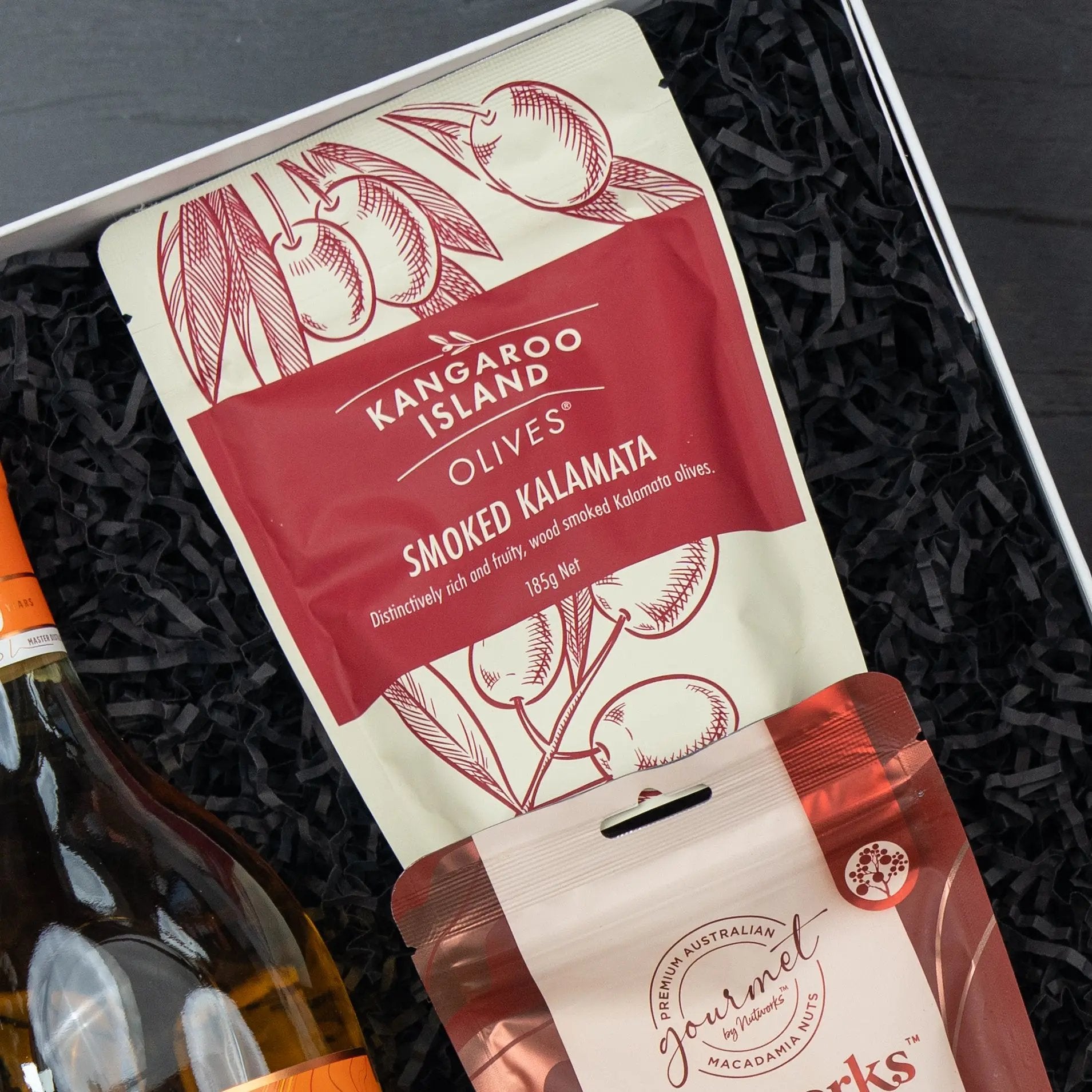 A close-up of The Hamper Boutique Co’s Glenmorangie Moments Hamper, showing a smoked kalamata olives packet resting on black shredded paper beside a bottle and another snack package inside the gift box.