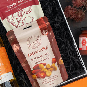 The Hamper Boutique Co’s Glenmorangie Moments Hamper features Salted Chilli Macadamias, smoked kalamata olives, a bottle of wine, red spread, and dried seed pods on a black shredded paper base.