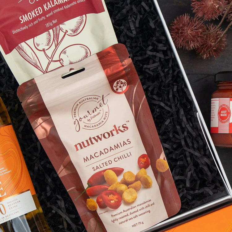 The Hamper Boutique Co’s Glenmorangie Moments Hamper features Salted Chilli Macadamias, smoked kalamata olives, a bottle of wine, red spread, and dried seed pods on a black shredded paper base.