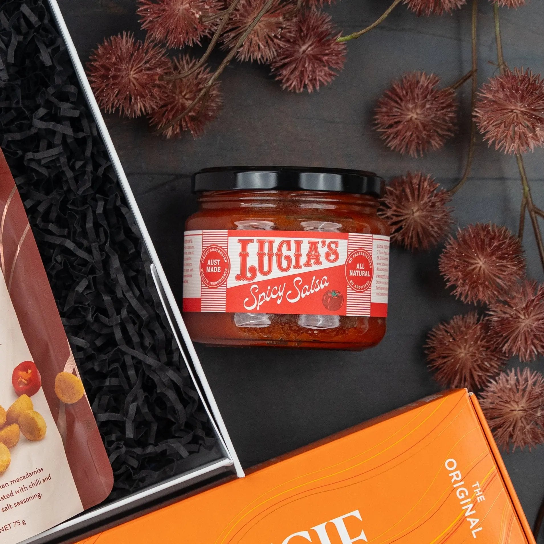 A Glenmorangie Moments Hamper by The Hamper Boutique Co rests on a dark surface, surrounded by decorative brown seed pods and partially visible food packages.