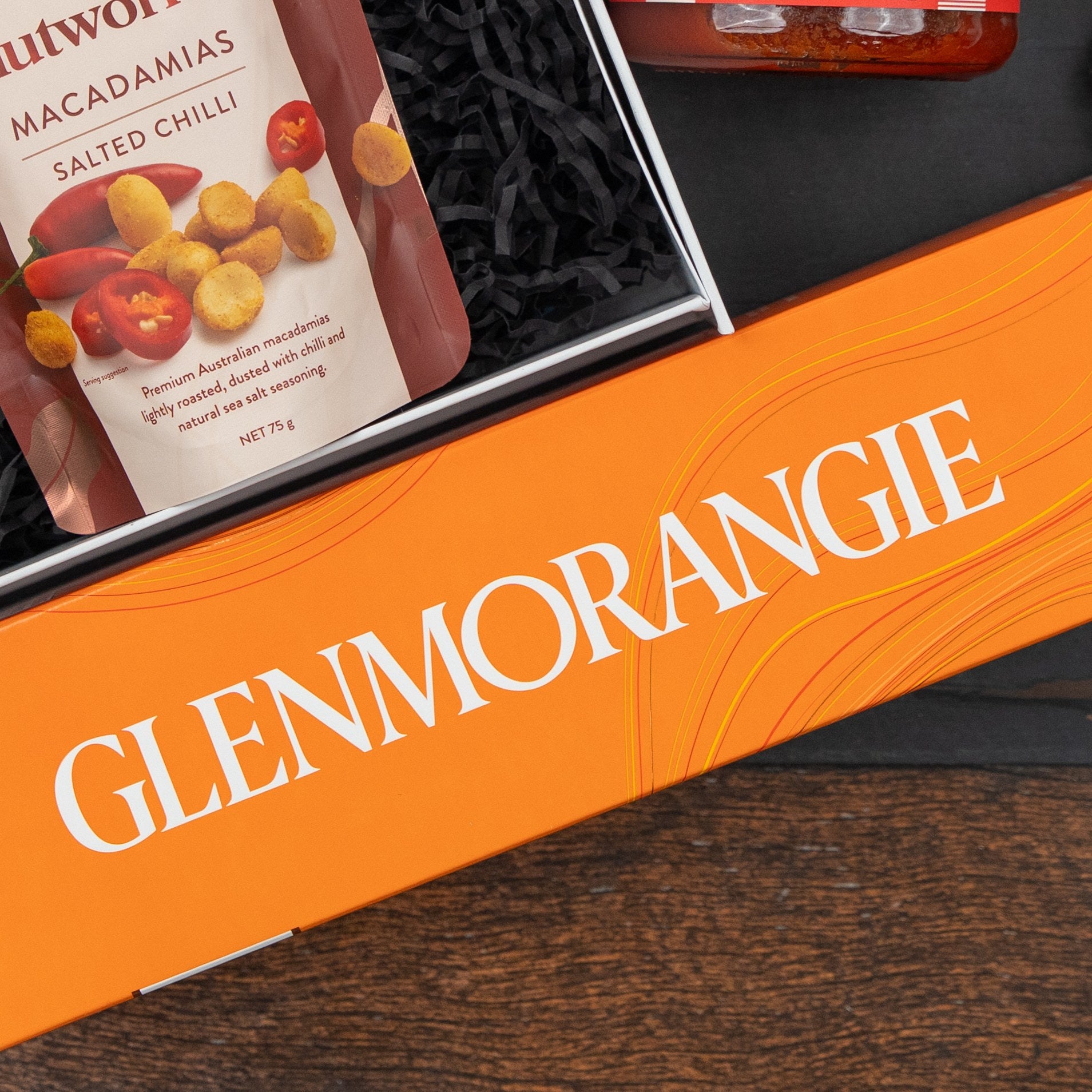 The Hamper Boutique Co's Glenmorangie Moments Hamper, featuring an orange box and Nutworks Macadamias Salted Chilli, is displayed on a dark surface with black shredded paper.