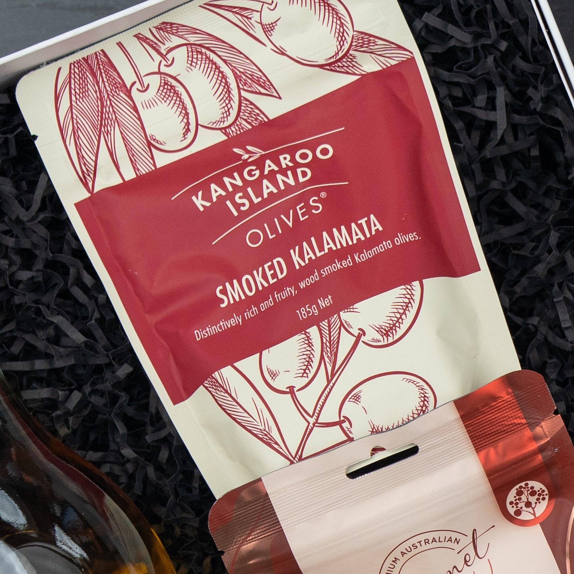 A Glenmorangie Moments Hamper from The Hamper Boutique Co rests on black shredded paper. The cream and red packaging features illustrated olive branches and contains a selection of items.