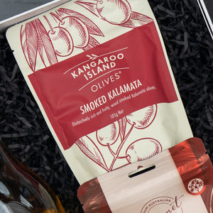 A Glenmorangie Moments Hamper from The Hamper Boutique Co rests on black shredded paper. The cream and red packaging features illustrated olive branches and contains a selection of items.