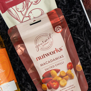 A Glenmorangie Moments Hamper from The Hamper Boutique Co lies on shredded black paper, with part of another snack package and a bottle nearby. The hamper packaging features an elegant design with sophisticated branding elements.