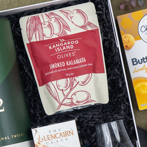 A Glenfiddich Connoisseur's Hamper by The Hamper Boutique Co is displayed in a gift box with black shredded paper, alongside partially visible green packaging and glassware.