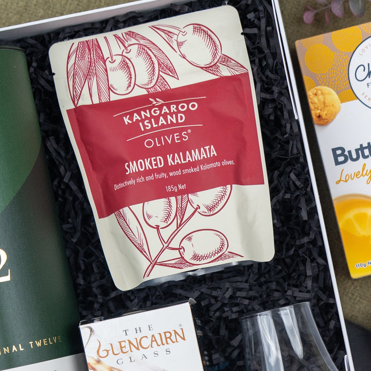 A Glenfiddich Connoisseur's Hamper by The Hamper Boutique Co is displayed in a gift box with black shredded paper, alongside partially visible green packaging and glassware.