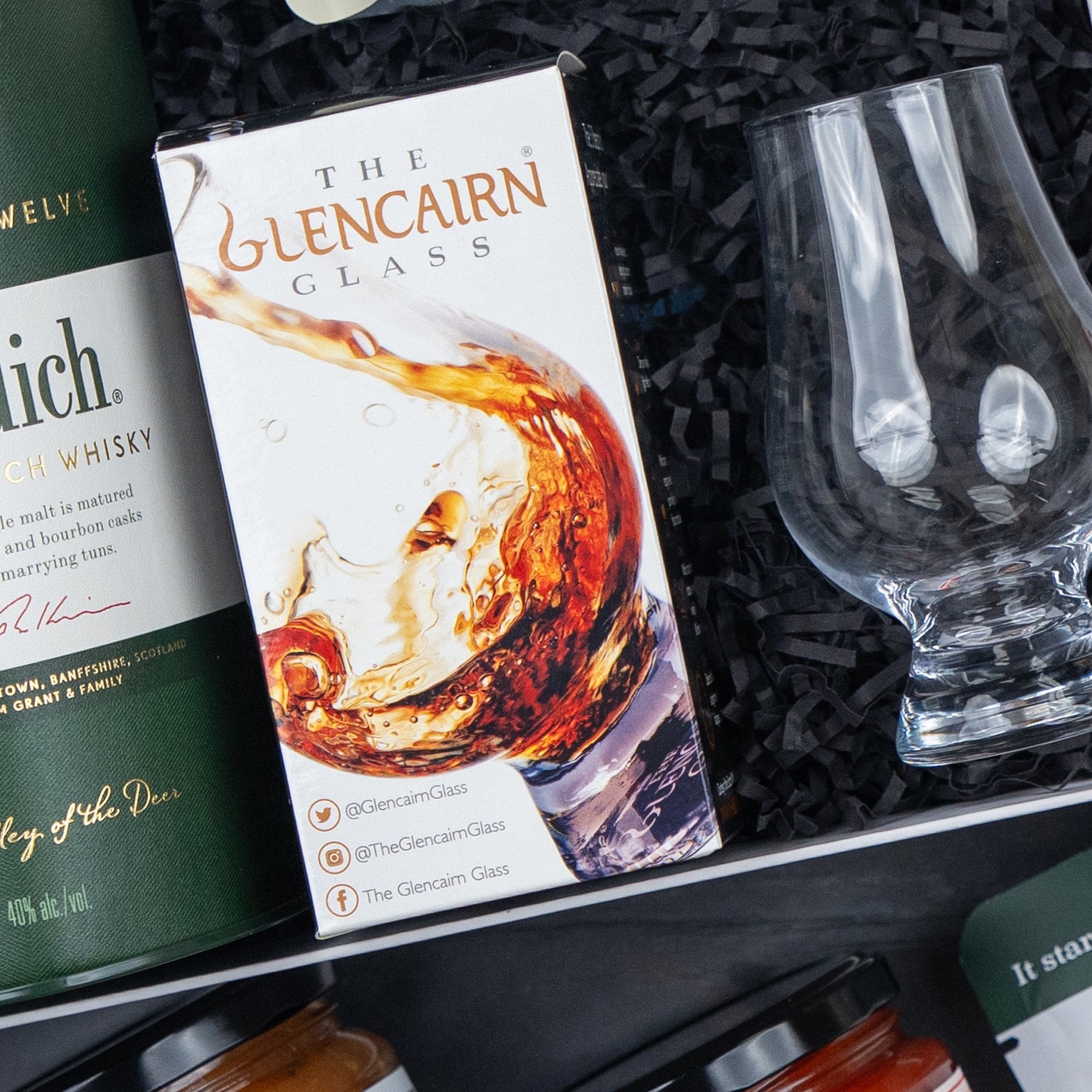 A Glenfiddich Connoisseur's Hamper by The Hamper Boutique Co, featuring a Glencairn Glass box with a whisky splash image, an empty glass, and a green whisky bottle on black shredded paper.