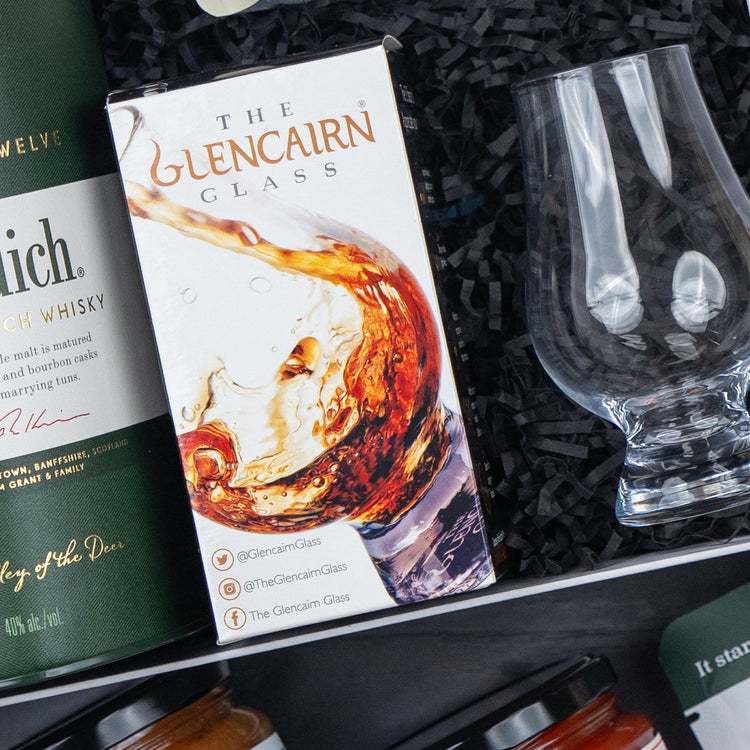 A Glenfiddich Connoisseur's Hamper by The Hamper Boutique Co, featuring a Glencairn Glass box with a whisky splash image, an empty glass, and a green whisky bottle on black shredded paper.