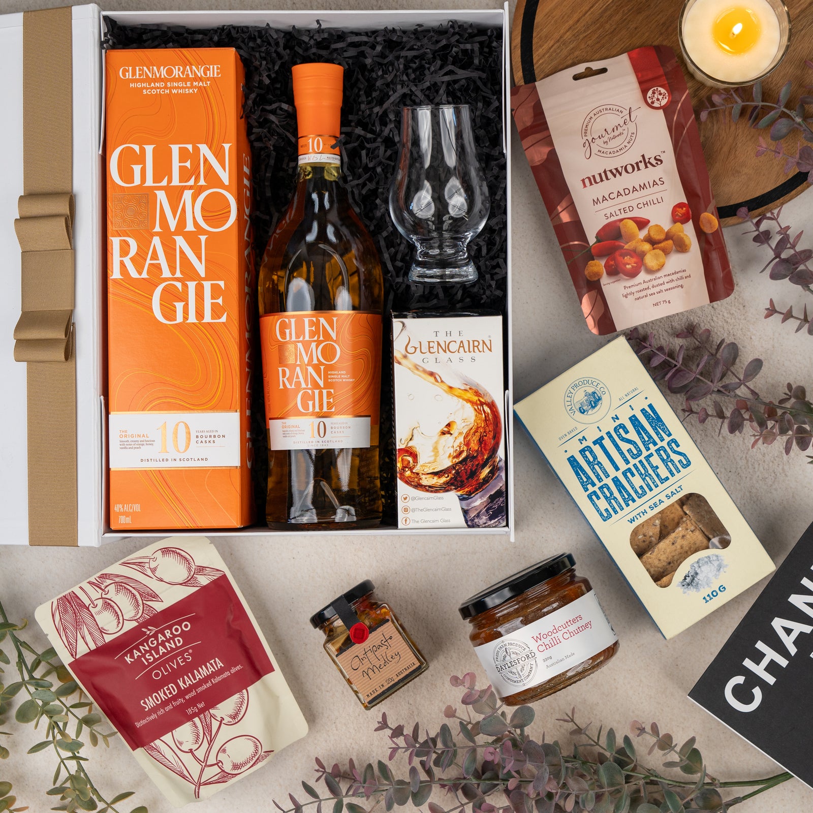 The Hamper Boutique Co’s Glenmorangie Discovery Hamper features Glenmorangie 10 whisky, a whisky glass, salted chilli macadamias, artisan crackers, smoked kalamata olives, quince paste, a candle, and eucalyptus leaves.