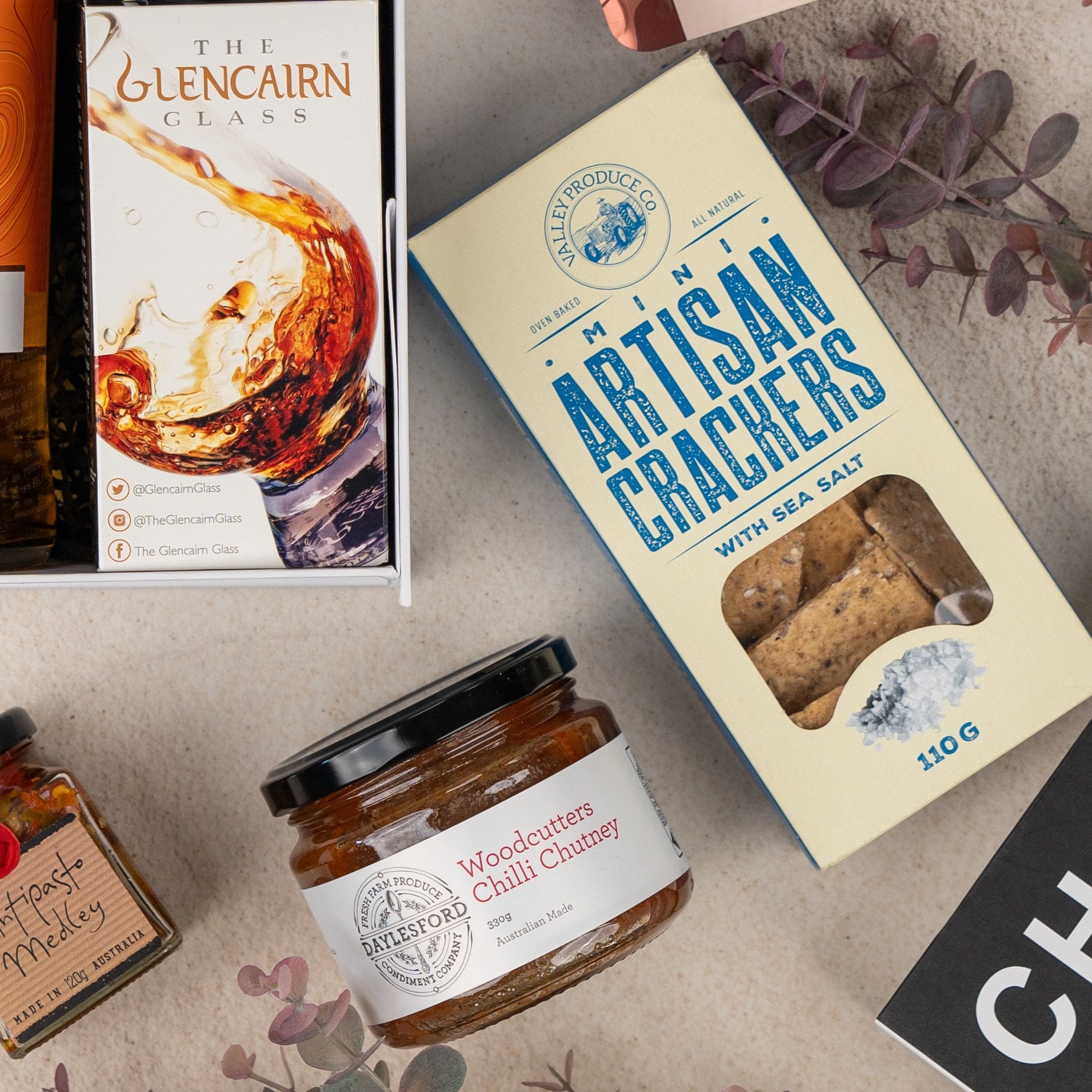 A flat lay of the Glenmorangie Discovery Hamper by The Hamper Boutique Co featuring a Glencairn whisky glass box, artisan crackers, Woodcutter’s Chilli Chutney, assorted preserves, and dried leaves on a beige background.