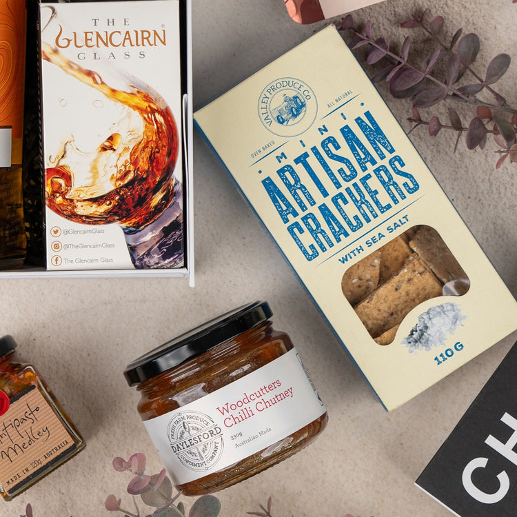 A flat lay of the Glenmorangie Discovery Hamper by The Hamper Boutique Co featuring a Glencairn whisky glass box, artisan crackers, Woodcutter’s Chilli Chutney, assorted preserves, and dried leaves on a beige background.