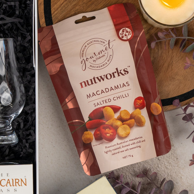 A Glenmorangie Discovery Hamper from The Hamper Boutique Co is displayed on a table with cocktail glasses, a candle, and scattered purple leaves. The packaging highlights the elegant branding of Glenmorangie.