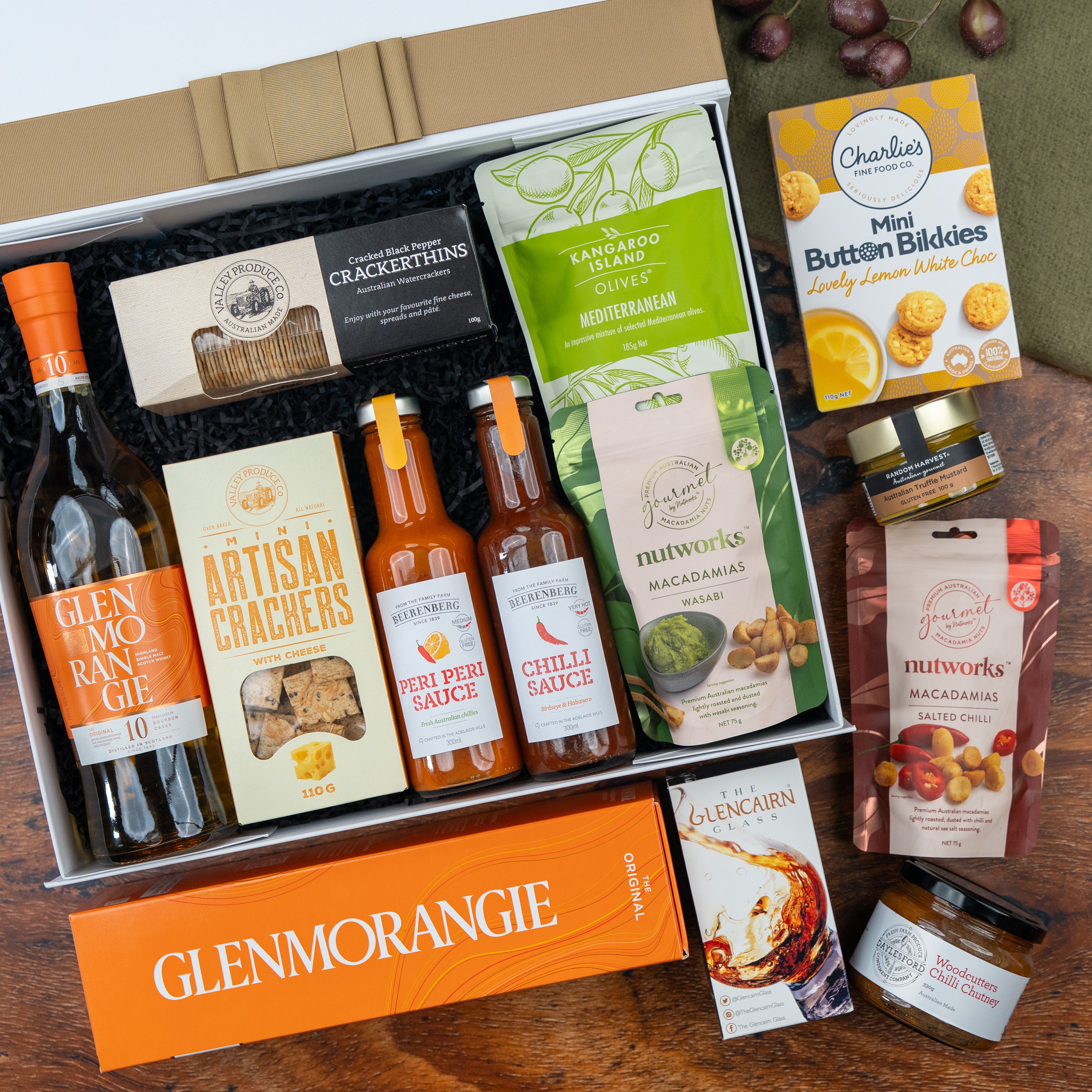 The Hamper Boutique Co’s Glenmorangie Premium Selection Hamper features Glenmorangie whisky, crackers, chilli sauces, macadamias, cookies, Kangaroo Island olives, quince paste and assorted snacks on a wooden display.
