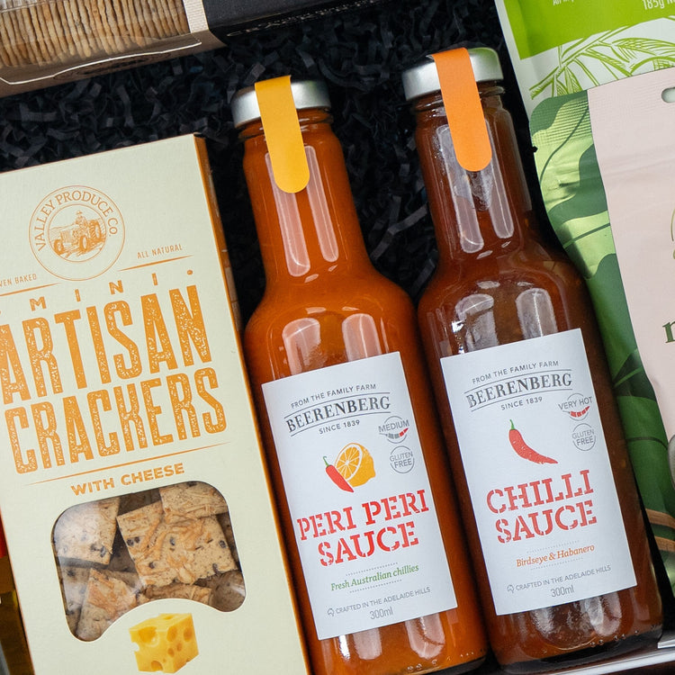 A close-up of The Hamper Boutique Co’s Glenmorangie Premium Selection Hamper, featuring mini artisan crackers with cheese and two bottles of Beerenberg Peri Peri Sauce and Chilli Sauce, with other gourmet items in the background.