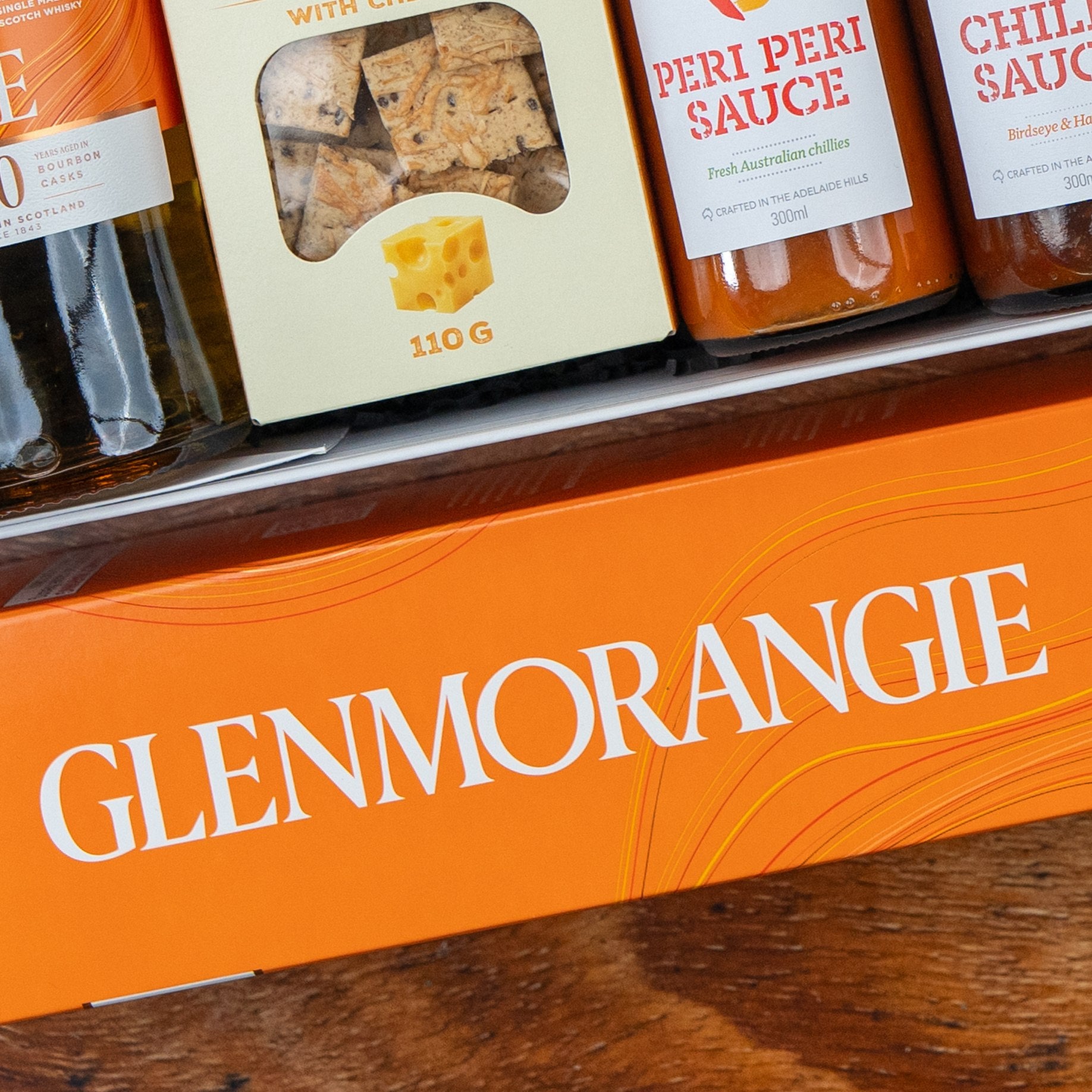 Close-up of The Hamper Boutique Co Glenmorangie Premium Selection Hamper box on a wooden surface, with bottles and snacks like peri peri sauce, chilli sauce, and crackers visible above the box.