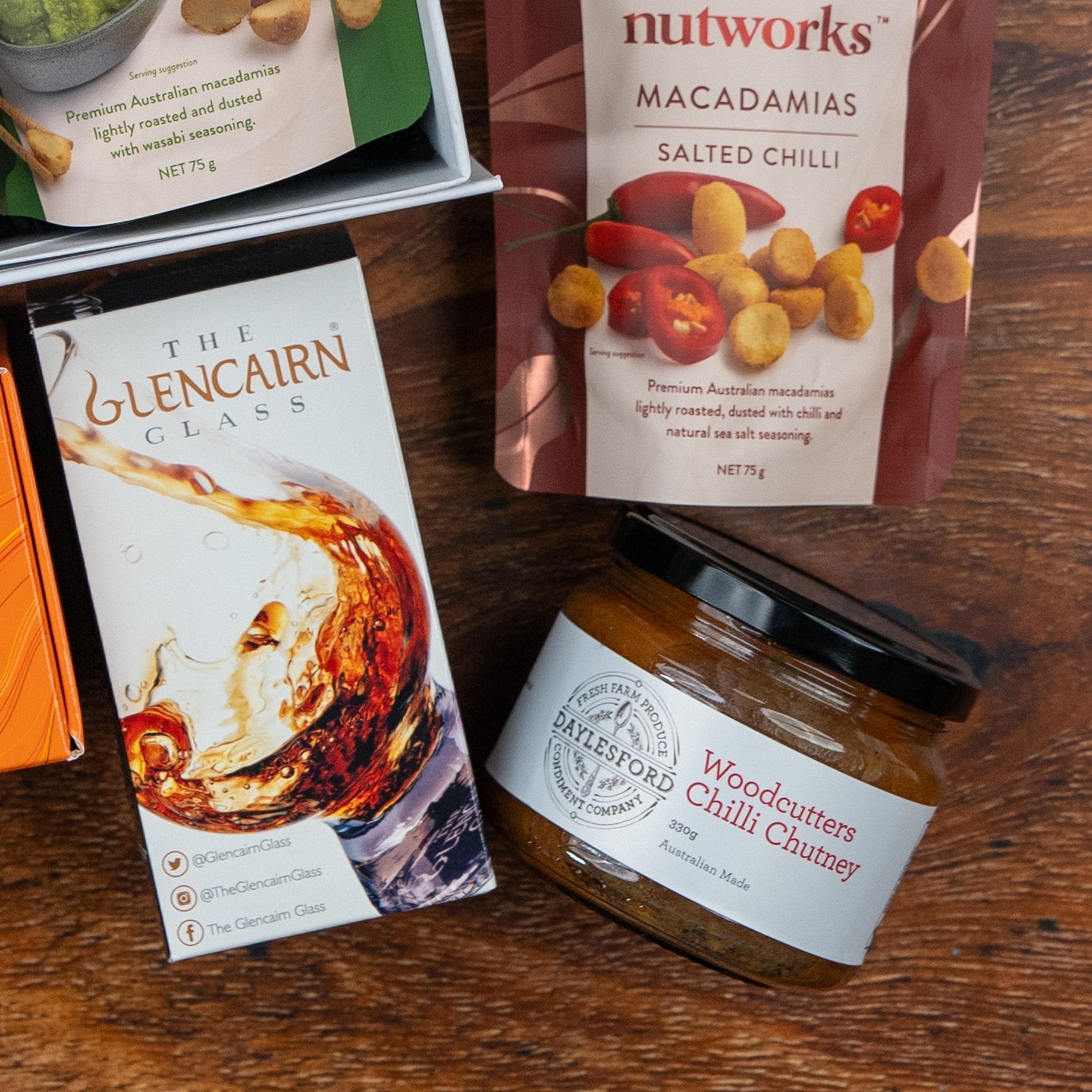 The Glenmorangie Premium Selection Hamper by The Hamper Boutique Co features a green bag of roasted macadamia nuts, a red bag of salted chilli macadamias, a Glencairn whisky glass box, and Woodcutters Chilli Chutney on a wooden surface.