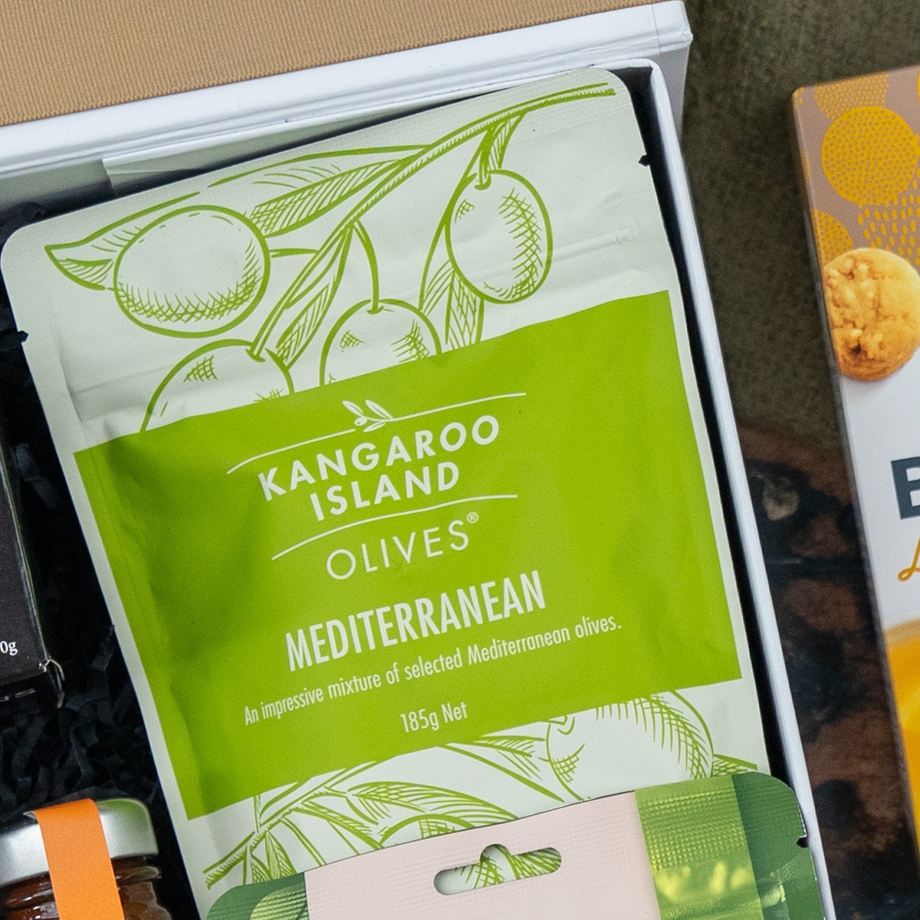 A green and white bag of Kangaroo Island Olives Mediterranean mix (185g) is included in The Hamper Boutique Co's Glenmorangie Premium Selection Hamper, with packaging featuring olive illustrations.