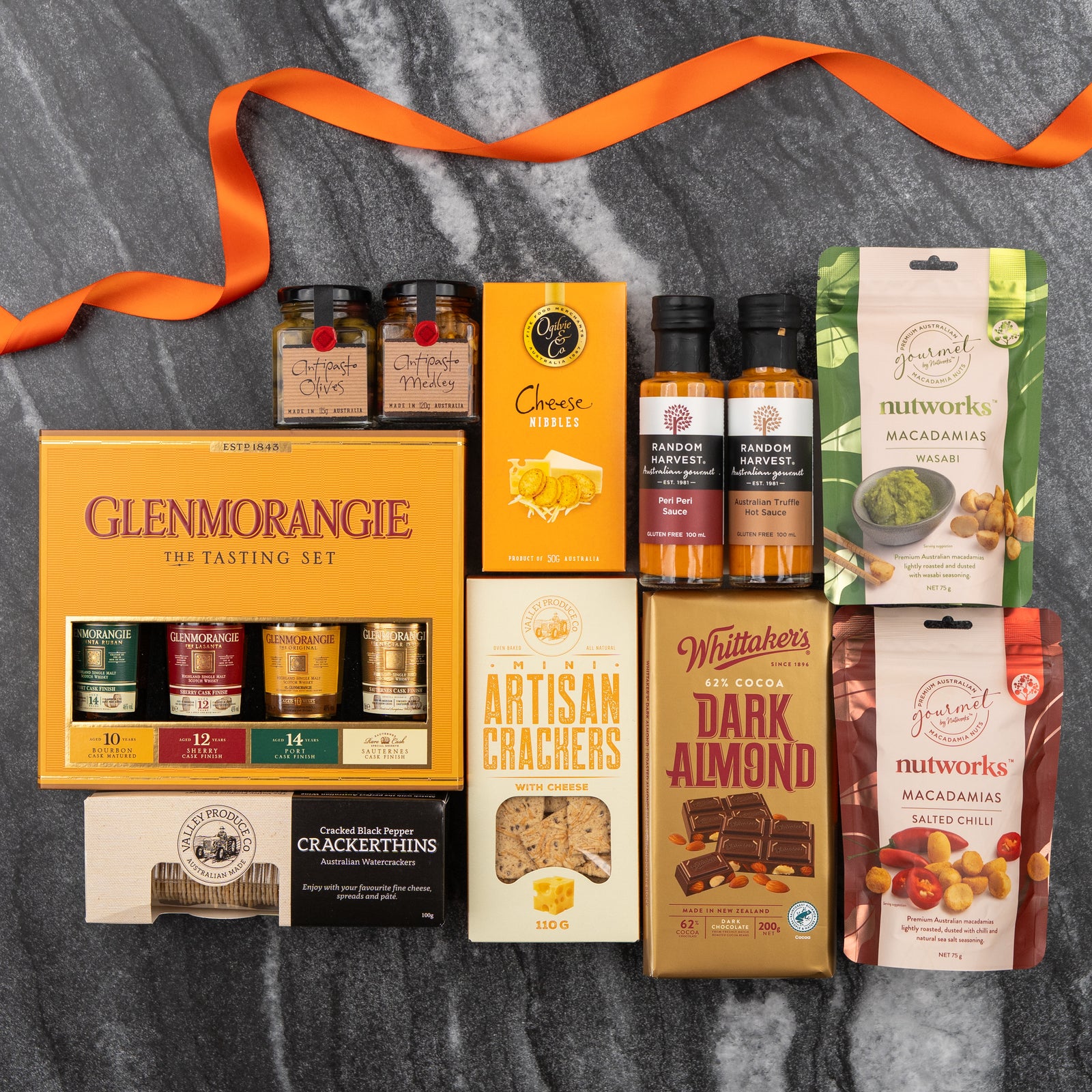 The Hamper Boutique Co's Glenmorangie Tasting Experience Hamper sits on marble, featuring Glenmorangie whisky samples, crackers, cheese nibbles, chutney, sauces, almond chocolate, and macadamia nuts—all adorned with an orange ribbon.