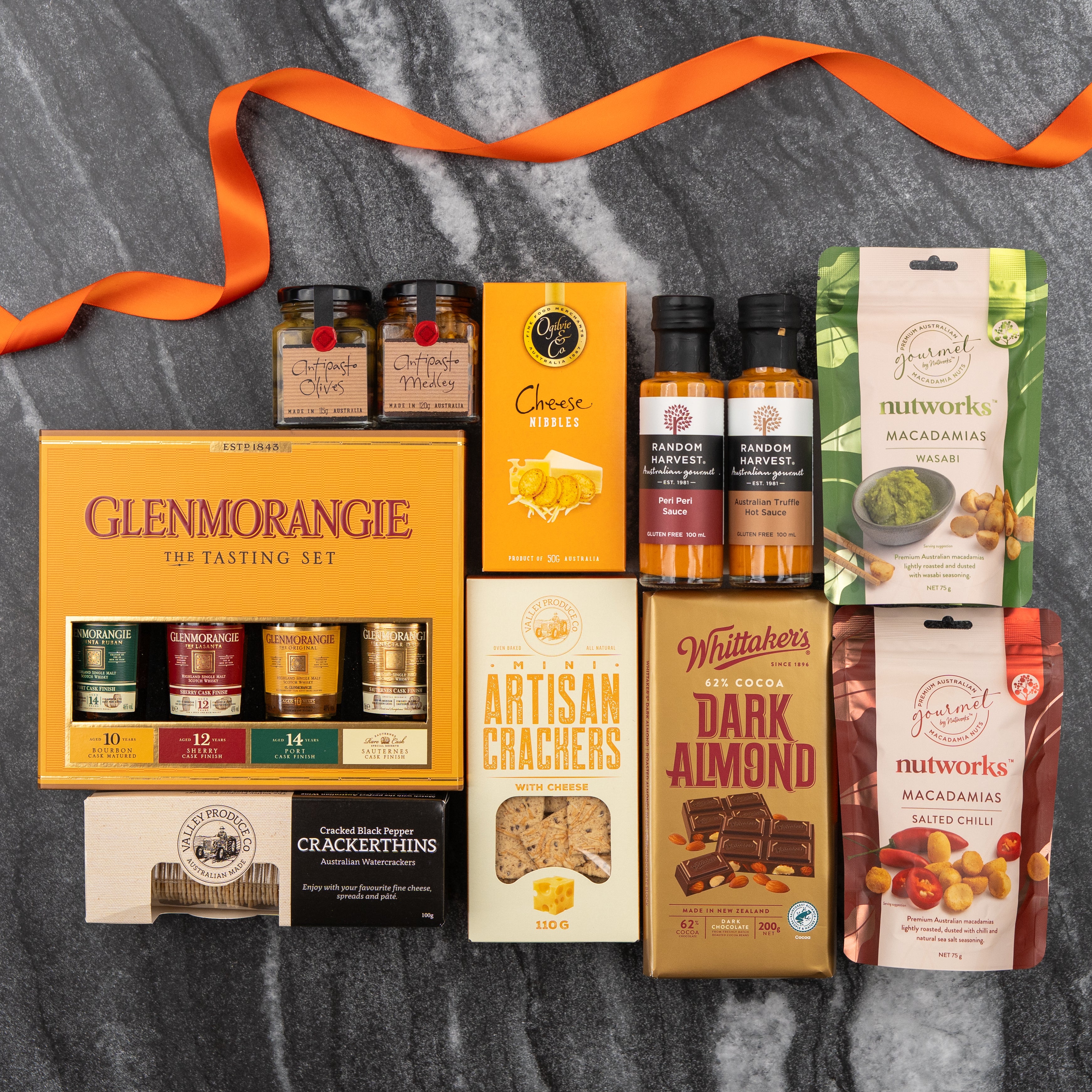 The Hamper Boutique Co's Glenmorangie Tasting Experience Hamper sits on marble, featuring Glenmorangie whisky samples, crackers, cheese nibbles, chutney, sauces, almond chocolate, and macadamia nuts—all adorned with an orange ribbon.