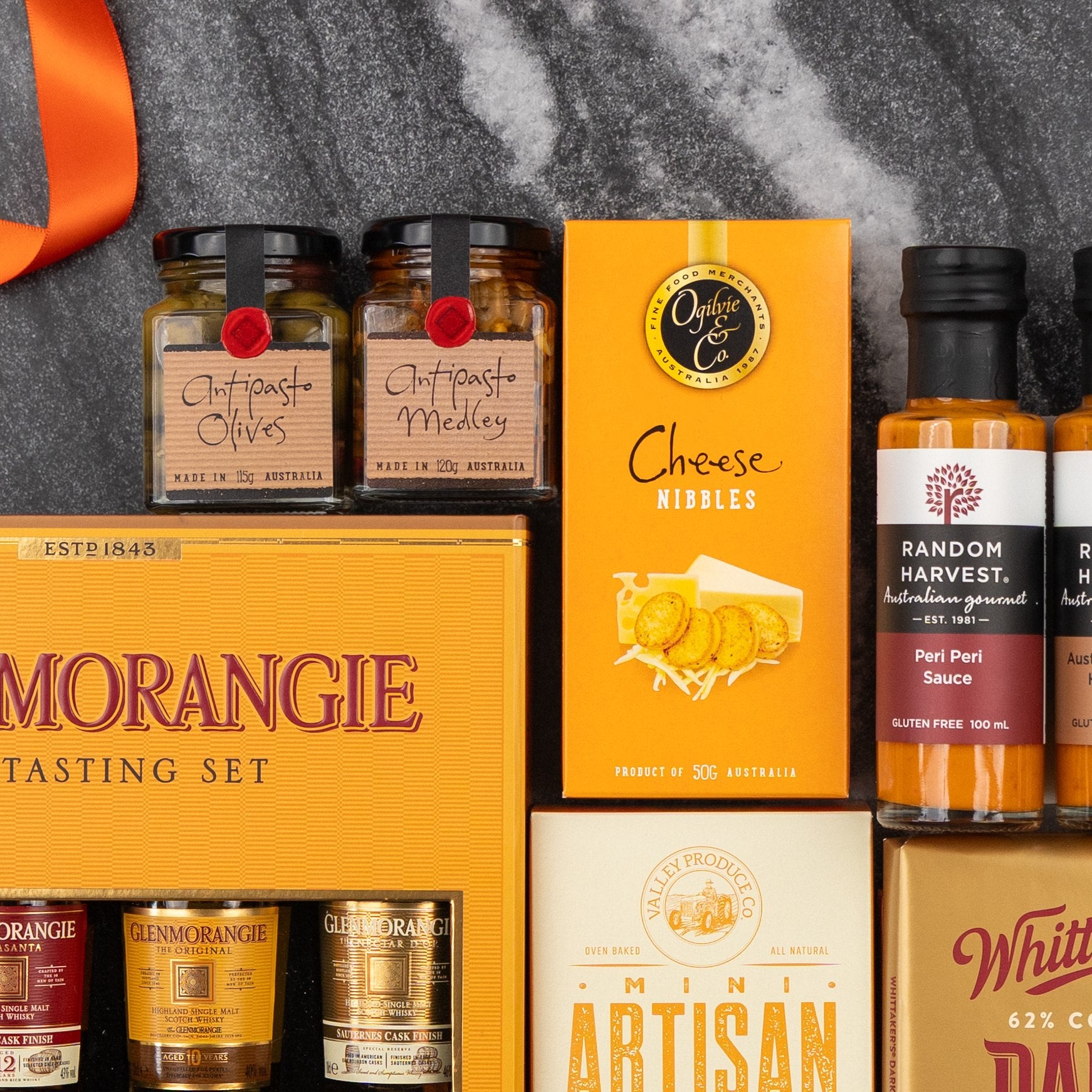 A flat lay of The Hamper Boutique Co Glenmorangie Tasting Experience Hamper on dark marble, featuring antipasto jars, cheese nibbles, artisan crackers, a whisky tasting set, and bottles of savoury and chocolate sauces.