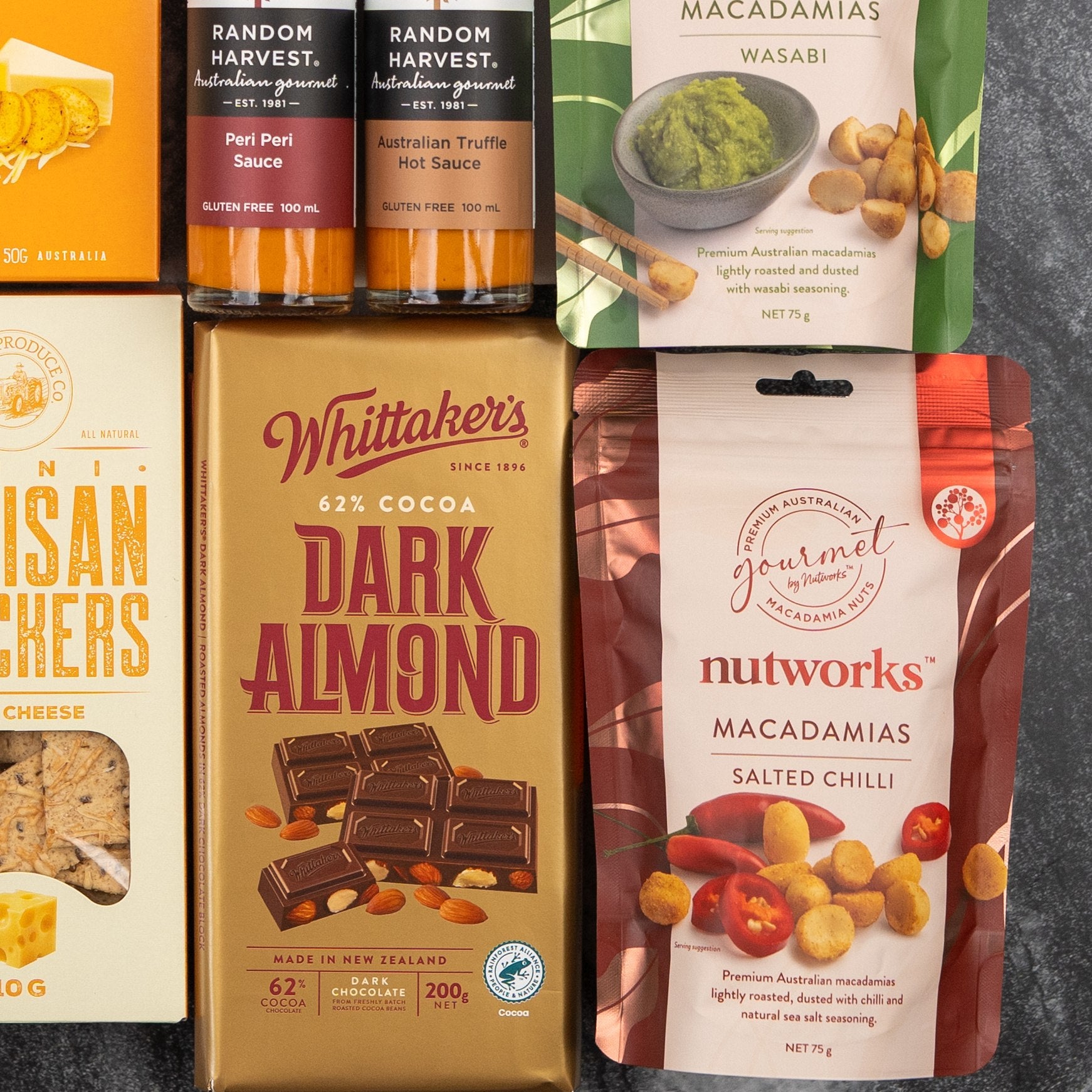 The Glenmorangie Tasting Experience Hamper by The Hamper Boutique Co features gourmet snacks—sauces, macadamias, crackers, and a Whittaker’s Dark Almond chocolate bar—in assorted colored packaging on a dark surface.
