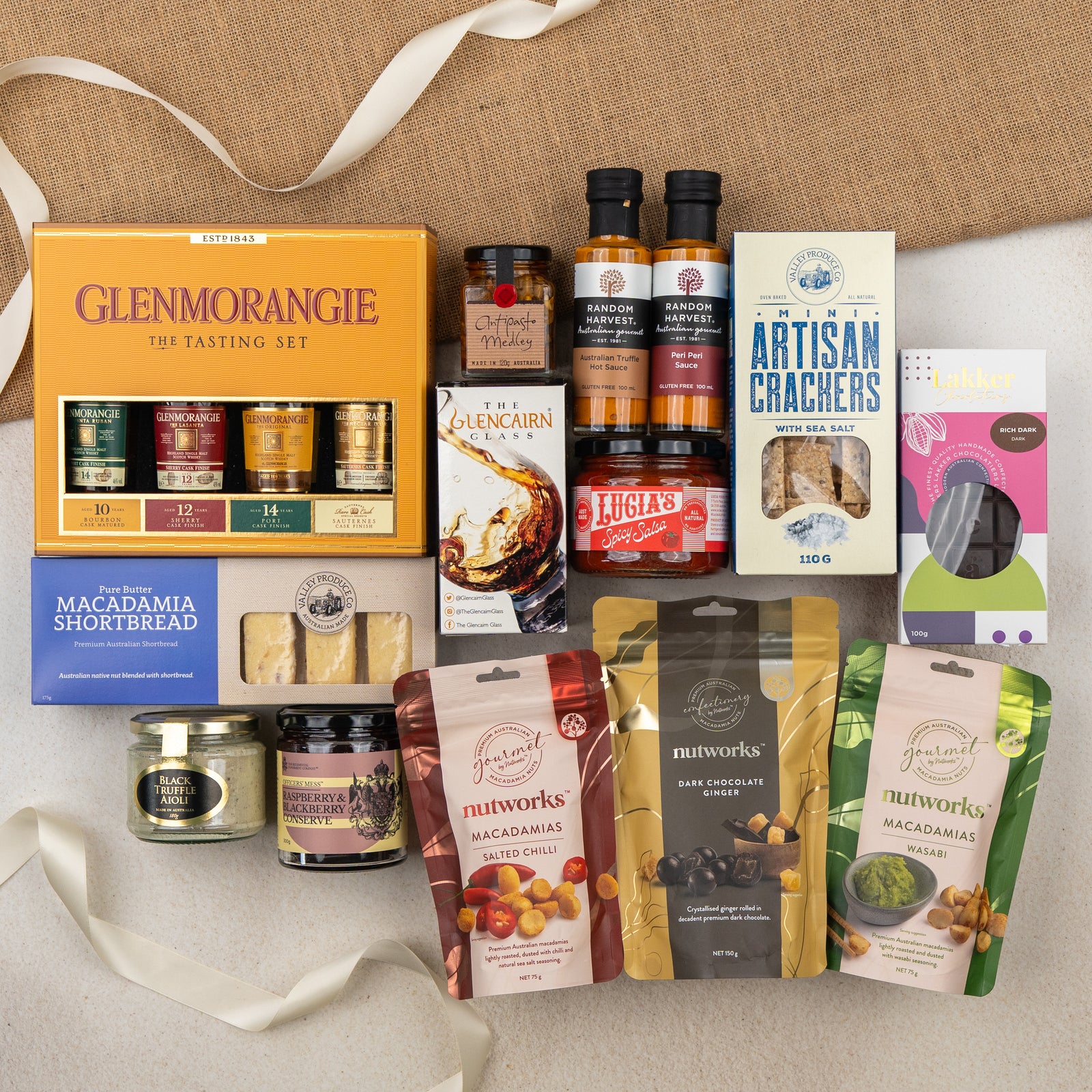 The Hamper Boutique Co’s Glenmorangie Ultimate Tasting Hamper features gourmet whisky, sauces, macadamia nuts, crackers, shortbread, chocolate, honey, and preserves—all elegantly arranged with ribbon and fabric accents on a beige surface.